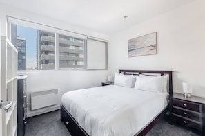 Ein modernes Hotelzimmer mit einem großen Bett, einem Nachttisch, einem Heizkörper, einem Fenster mit Blick auf Gebäude und einem Bild an der Wand.