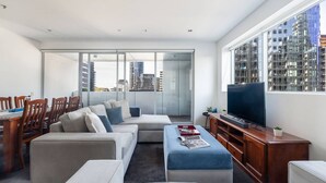 A modern living room with a sofa set, a dining table with chairs, a TV, and a large window with a city view. - Urban Calm Comfortable Base with City Views and Gym (South Yarra)