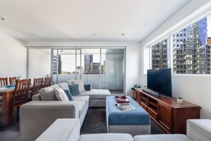 A modern living room with a sofa set, a dining table with chairs, a TV, and a large window with a city view. - Urban Calm Comfortable Base with City Views and Gym (South Yarra)