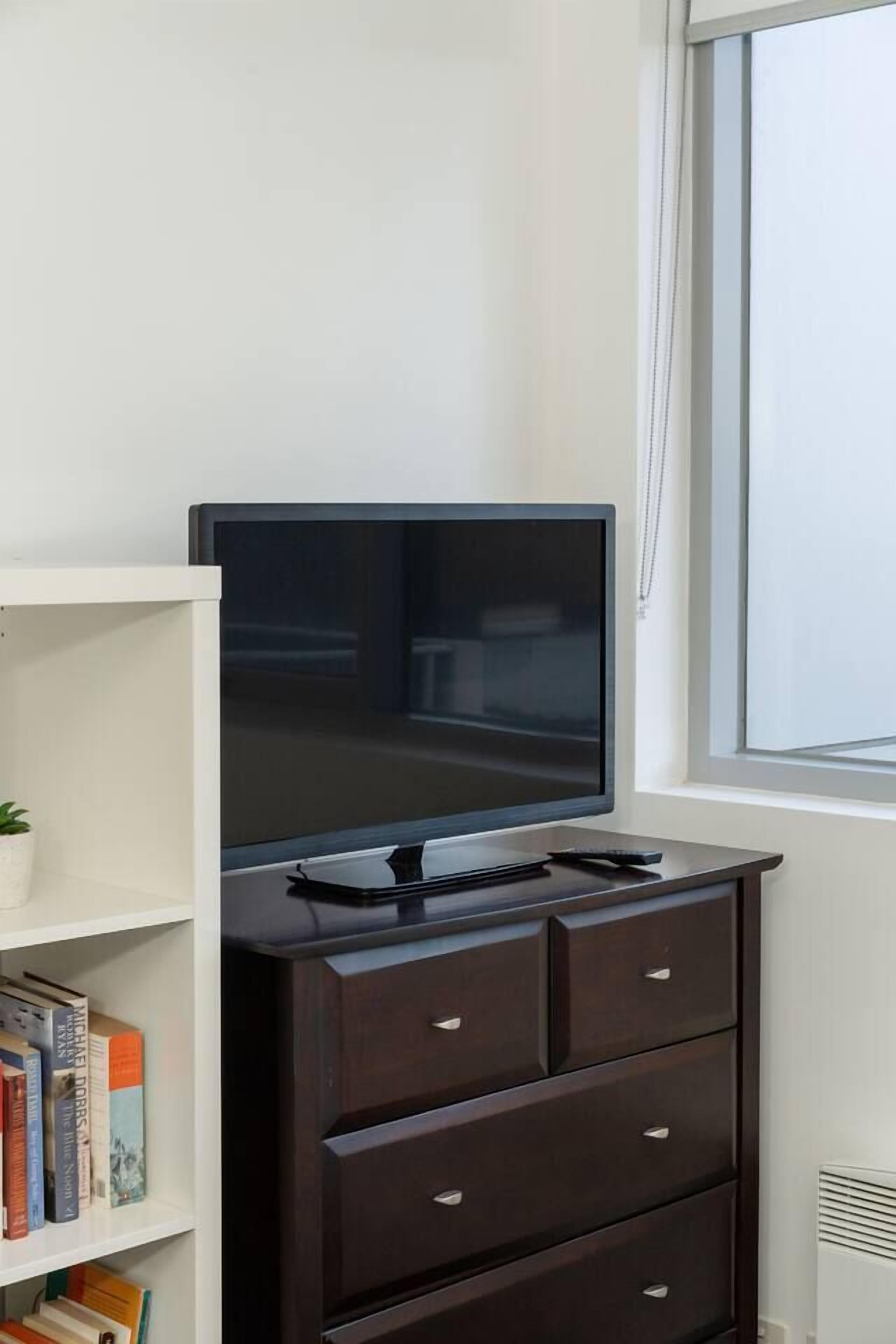 A flat-screen TV on a dark wooden dresser, a white bookshelf with books, and a window with blinds.