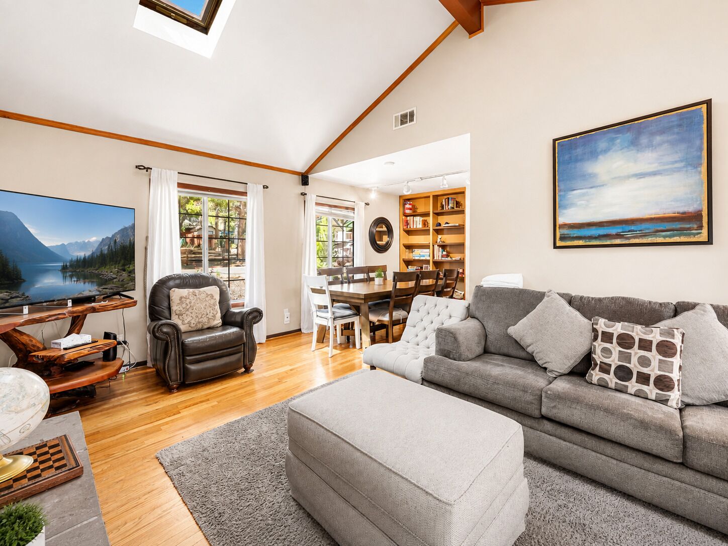 A spacious living area with a large flat-screen TV, a leather armchair, a grey sofa, and a dining table with chairs.