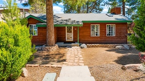 A wooden cabin with a green roof, a stone pathway leading to the entrance, and a large tree in front.