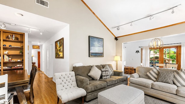 A spacious living room with a wooden dining table, two sofas, a grey ottoman, and a bookshelf.