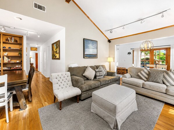 A spacious living room with a wooden dining table, two sofas, a grey ottoman, and a bookshelf.