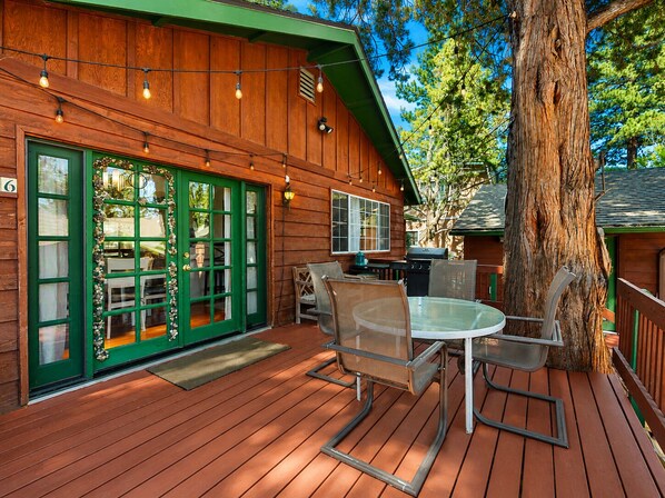 Una terraza de madera con mobiliario exterior, una parrilla y un gran árbol.