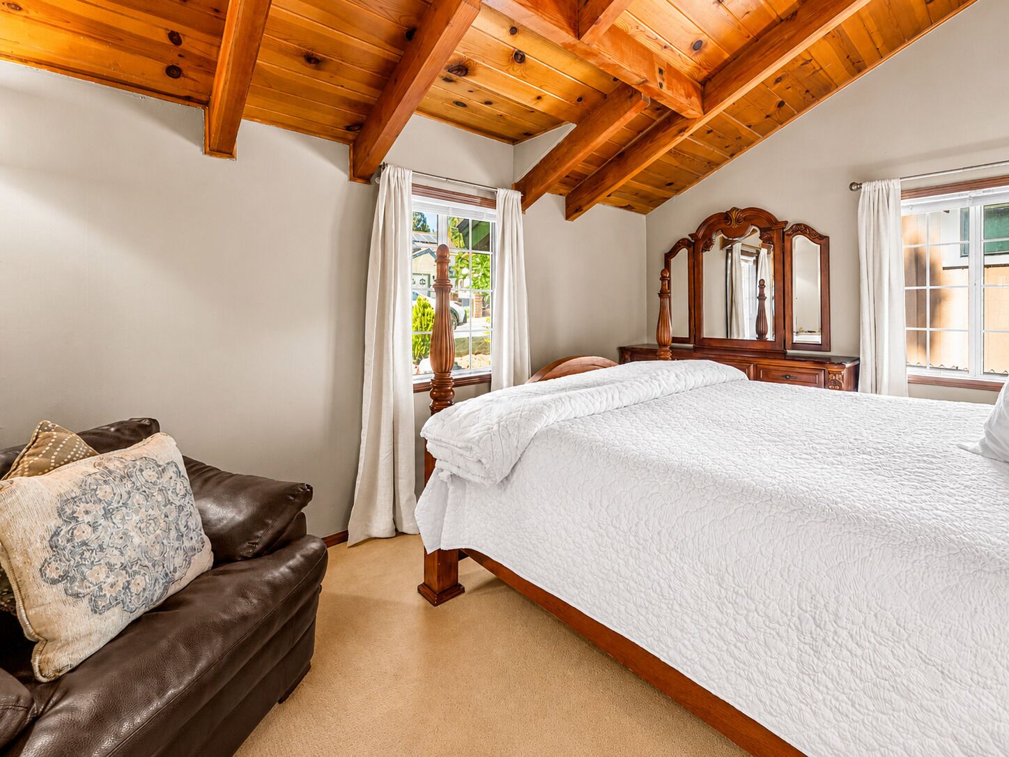 A bedroom with a large bed, a wooden ceiling, a window with curtains, and a brown leather sofa with a patterned pillow.