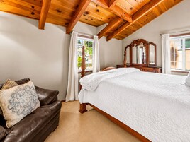 Un dormitorio con una cama grande, techo de madera, ventana con cortinas y un sofá de cuero marrón con un cojín estampado.