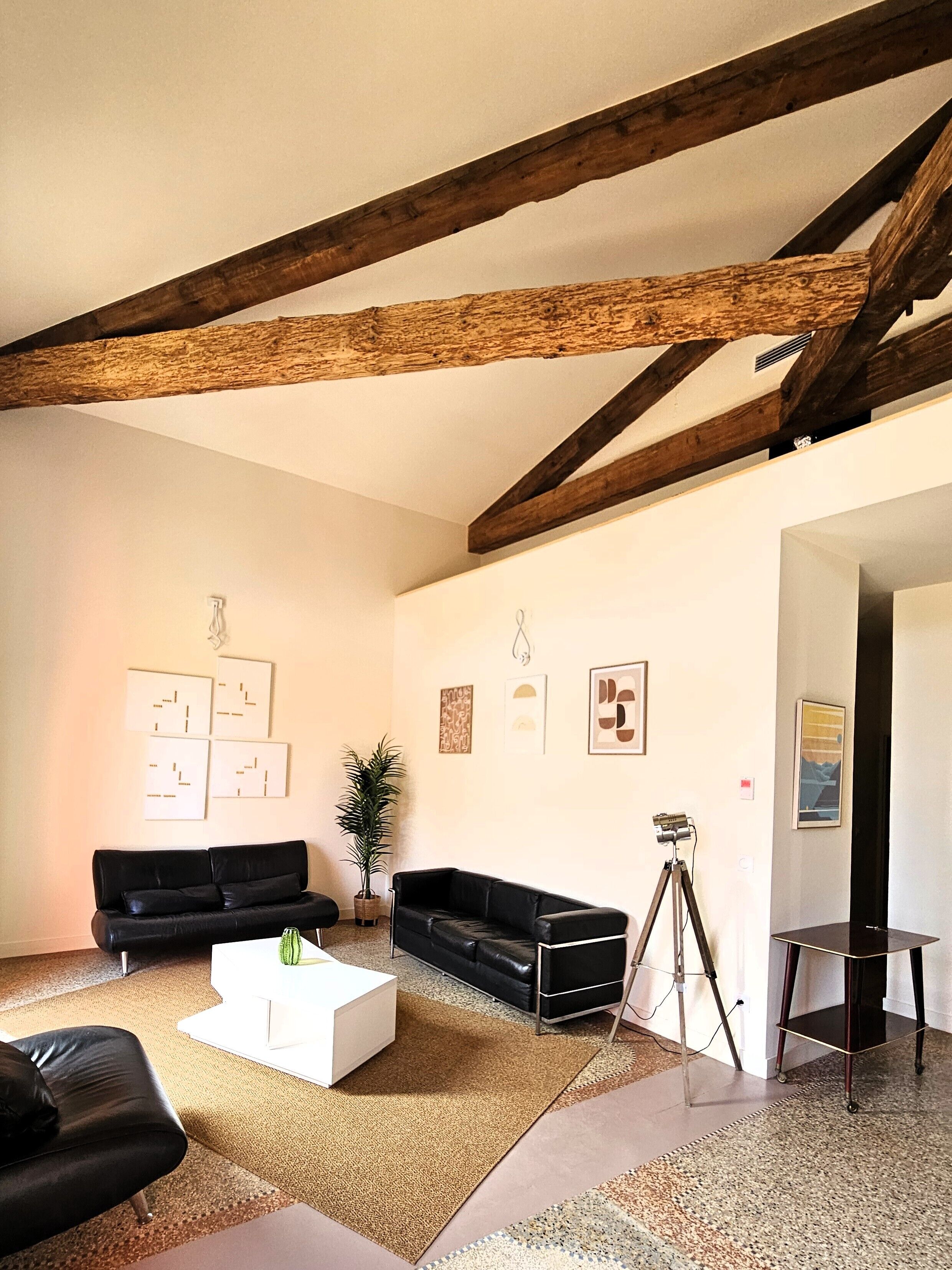 Sala de estar con vigas de madera a la vista, un sofá de cuero negro, una mesa de centro blanca y un trípode.