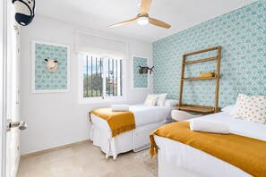 Une chambre avec deux lits, une fenêtre avec des barreaux, un ventilateur de plafond et un mur recouvert d’un papier peint à motifs.