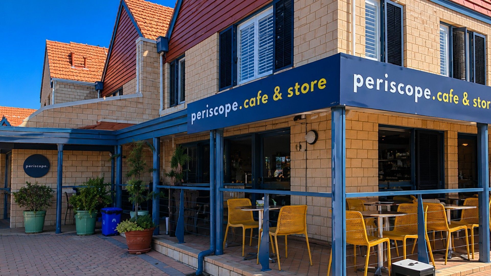 An outdoor cafe with yellow chairs and tables, a blue awning, and a sign that reads 'periscope cafe & store'.