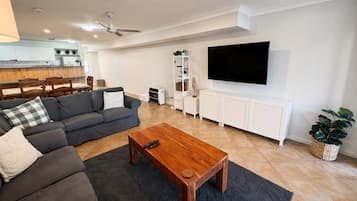 A modern living room with a grey sofa, a wooden coffee table, a TV on the wall, and a kitchen in the background.