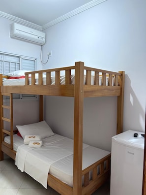 Una cama litera con estructura de madera, un colchón blanco y una mesita de noche blanca con un control remoto.