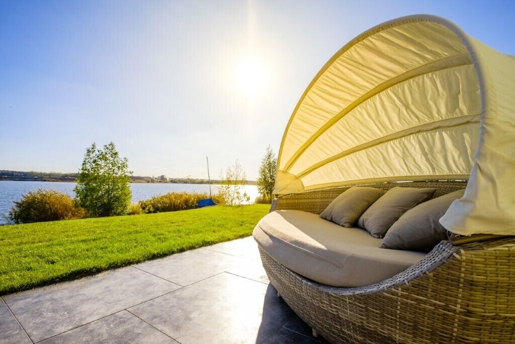 Ein Korbensofa im Freien mit Baldachin, platziert auf einer Terrasse mit Blick auf einen See und Grünflächen.