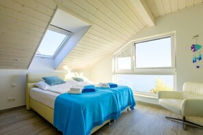 A bedroom with a sloped ceiling, a bed with blue bedding, a white chair, and a view of the sea through a window.