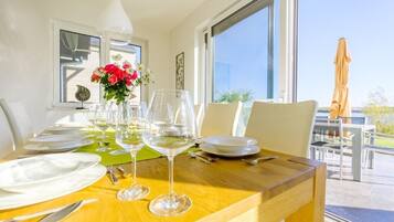 Mesa de comedor dispuesta con platos blancos, cubiertos y copas de vino, con un jarrón de flores y vistas al exterior.