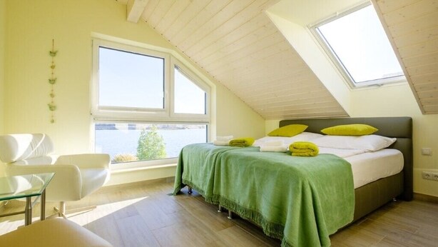 Un dormitorio con techo inclinado, una cama con ropa de cama verde y blanca, una ventana y vistas al exterior.