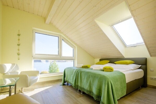 A bedroom with a sloped ceiling, a bed with green and white bedding, a window, and a view of the outdoors.