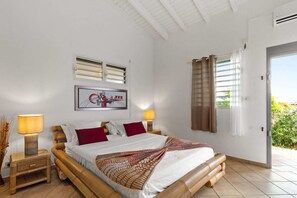 Een slaapkamer met een bed, twee bedlampen, een raam met gordijnen, een aan de muur hangend schilderij en een deur naar buiten.