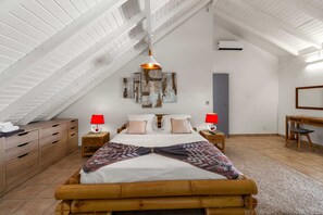 A bedroom with a sloped ceiling, a bed with a patterned blanket, two bedside tables with lamps, a wall-mounted air conditioner, and a desk with a chair. - Villa Palmera - Four Bedroom Villa, Sleeps 8 (Saint-François)