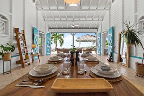 A dining area with a view of the sea, featuring a long table set for a meal, a ceiling fan, and a tropical plant. - Villa Zouina - Three Bedroom Villa, Sleeps 7 (Le Carbet)