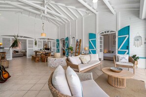 A spacious living area with a high ceiling, open doors, and a mix of seating arrangements including a sofa, chairs, and a dining table.