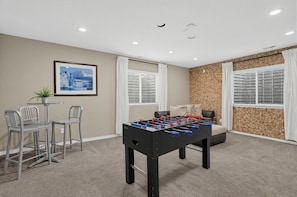 Una habitación con mesa de futbolín, sofá, mesa de comedor con sillas y un cuadro en la pared.