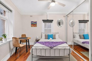 A bedroom with a bed, a chair, a table, a ceiling fan, and a window. - Roslyn Studio Between Harbour Calm and City Energy (Potts Point)