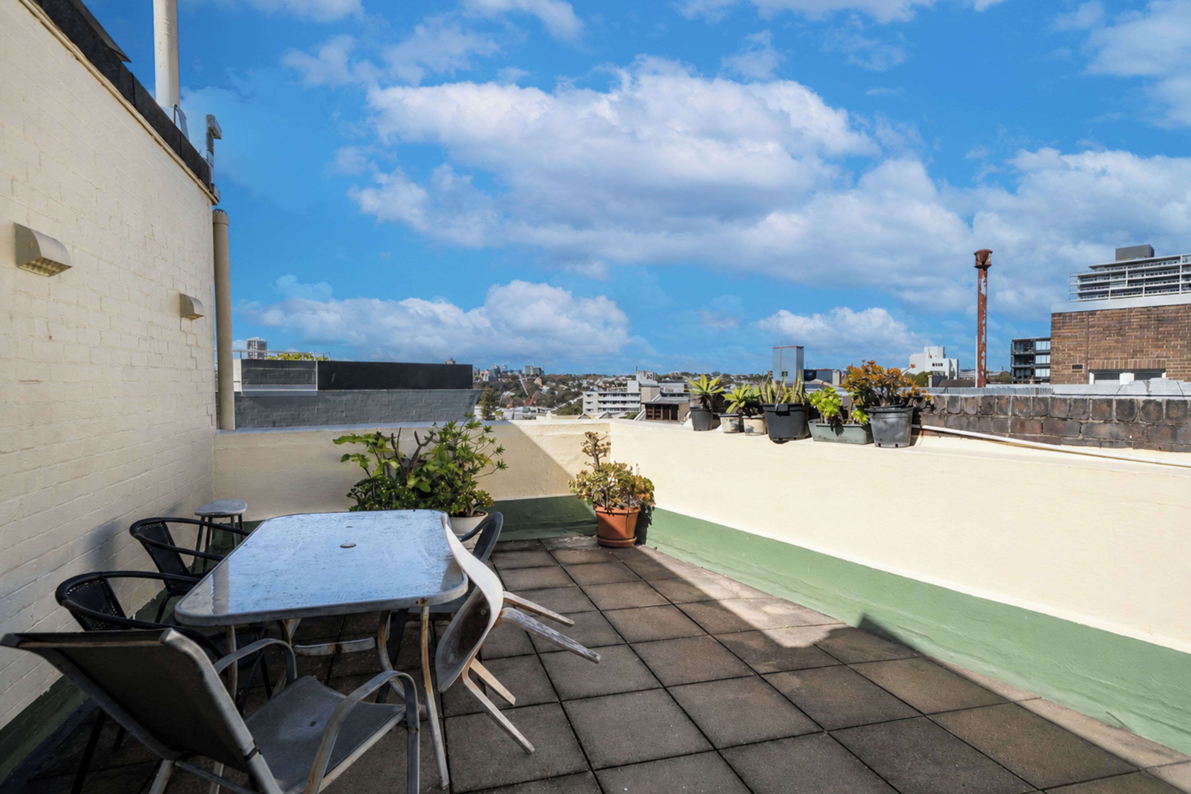 A rooftop terrace with a table and chairs, potted plants, and a view of the cityscape.