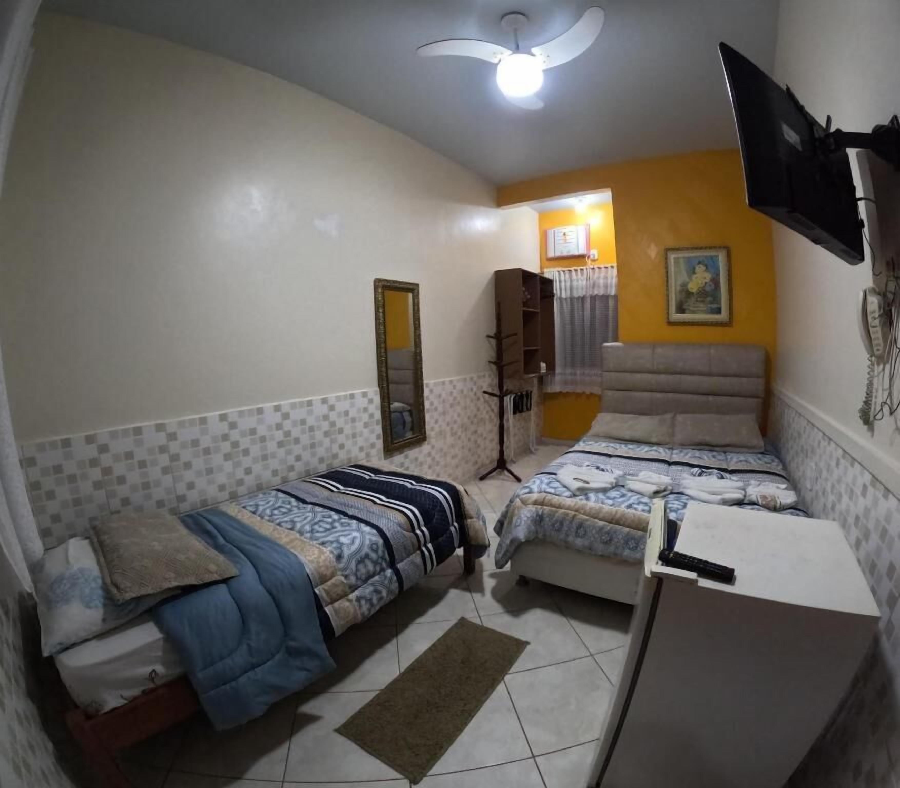 Quarto com duas camas, ventilador de teto, TV na parede e uma escrivaninha.