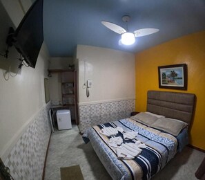 A bedroom with a bed, a ceiling fan, a television mounted on the wall, and a small refrigerator.