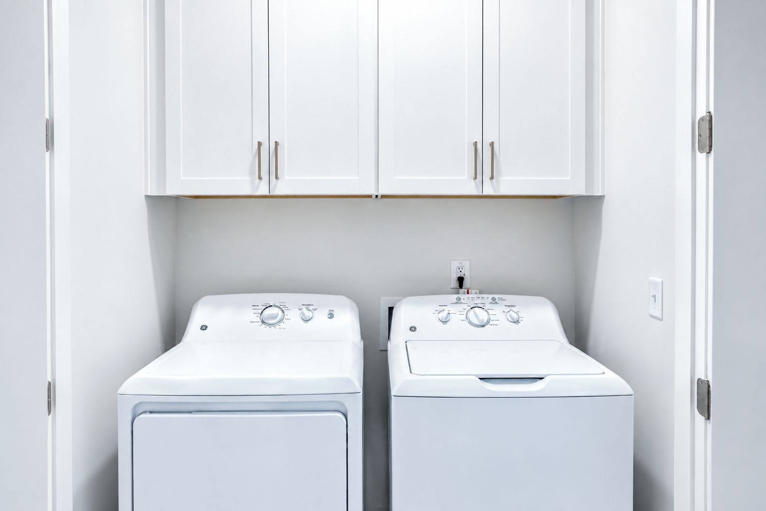 A laundry room with two white front-loading washing machines and white cabinets above.