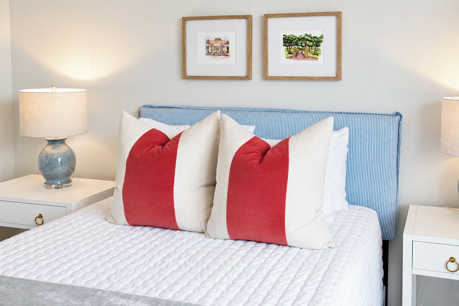 Una cama bien hecha con ropa blanca y almohadas rojas y blancas, flanqueada por dos mesitas de noche con lámparas y cuadros en la pared.