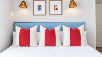 Uma cama com cobertura branca acolchoada, cabeceira listrada em azul e branco e almofadas vermelhas e brancas.