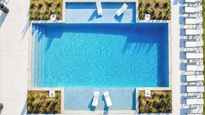 En rektangulär pool med klart, blått vatten, omgiven av vita solstolar och små, gröna växter.