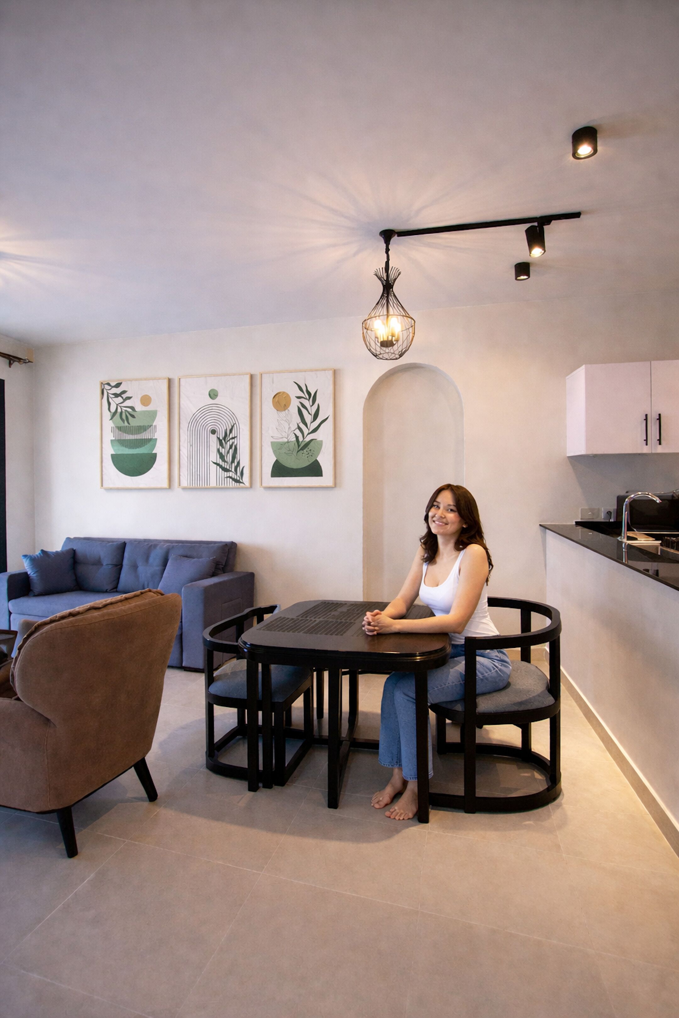 A modern living room with a sofa, a round table, and chairs. There are wall-mounted artworks and a ceiling light fixture.