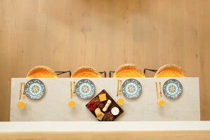 A dining setup with a white tablecloth, four plates with intricate designs, golden cutlery, and baskets of bread.