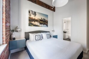 Ein Schlafzimmer mit Bett, Nachttischen, einem an der Wand hängenden Bild und einem durch eine Glastür einsehbaren Badezimmer.