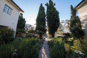 Ein Gartenweg führt zu einem Gebäude mit weißer Fassade, umgeben von hohen Bäumen und verschiedenen Sträuchern.