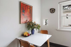 Ein Essbereich mit einem weißen Tisch, zwei Stühlen, einer Blumenvase, einer Obstschale, einem Gemälde, einer Uhr und einem Fenster mit Blick auf Gebäude.