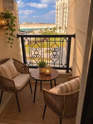 A balcony with wicker furniture, a small table, and a view of a beach and buildings.