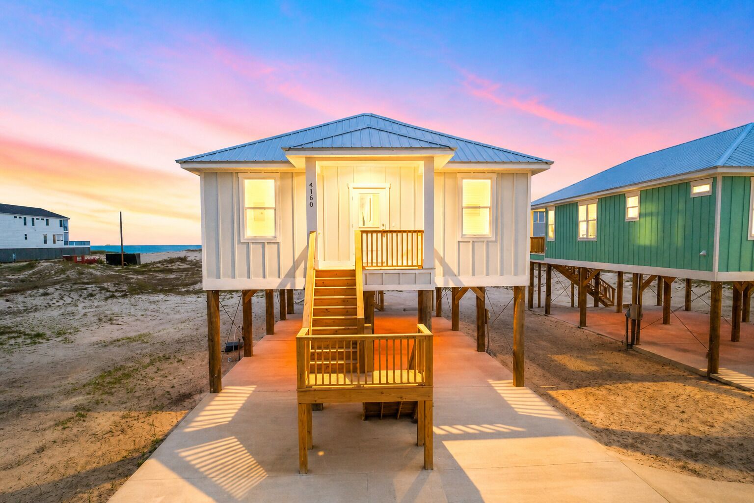 Ein Strandhaus mit einer Holztreppe zum Eingang, einem Betonweg und einem Sandstrand im Hintergrund.