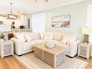 A living room with a beige sectional sofa, a wooden coffee table, and a painting of a beach scene. - "How You Dune"
  Serene 3-bedroom house with GULF views in Fort Morgan! (Gulf Shores)