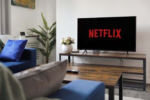 A living room with a flat-screen TV displaying 'NETFLIX' on the screen, a blue sofa, a coffee table, and a potted plant. - Cozy 2BR Home Sleeps 5 with WiFi by Stay With Us (Platt Bridge)