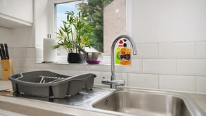 A kitchen with a stainless steel sink, a dish rack, a potted plant, and a window with a view of greenery. - Cozy 2BR Home Sleeps 5 with WiFi by Stay With Us (Platt Bridge)
