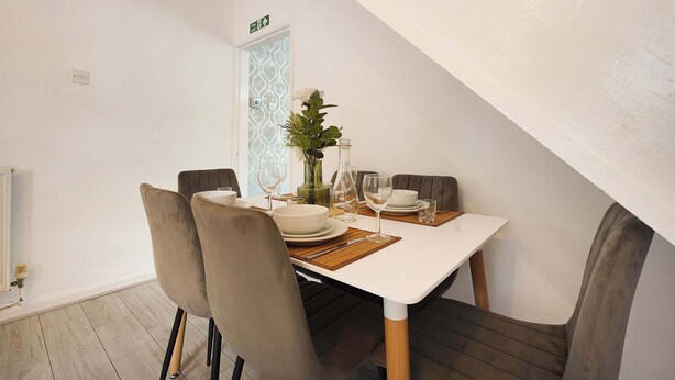 A dining area with a white table, four chairs, and a vase with flowers. - Cozy 2BR Home Sleeps 5 with WiFi by Stay With Us (Platt Bridge)