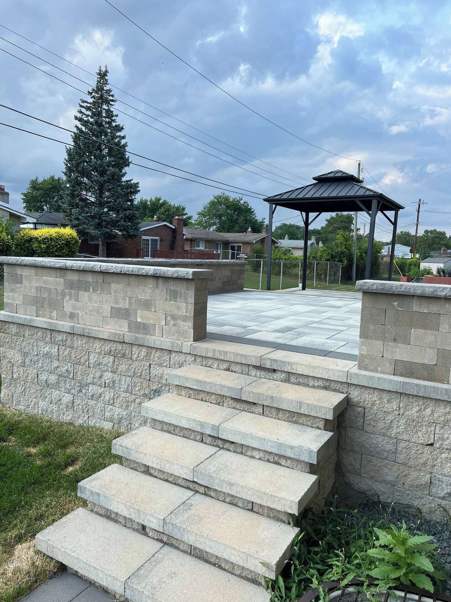 A stone staircase leading to a covered outdoor area with a metal roof, surrounded by a low stone wall and greenery.