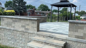 A stone staircase leading to a covered outdoor area with a metal roof, surrounded by a low stone wall and greenery.