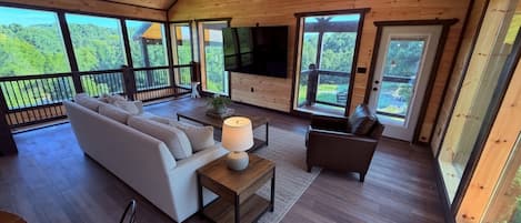 A spacious living room with a sofa, armchair, and a TV, large windows offering a view of the outdoors.