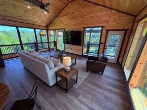Ein geräumiges Wohnzimmer mit Sofa, Sessel und Fernseher, große Fenster mit Blick ins Freie.