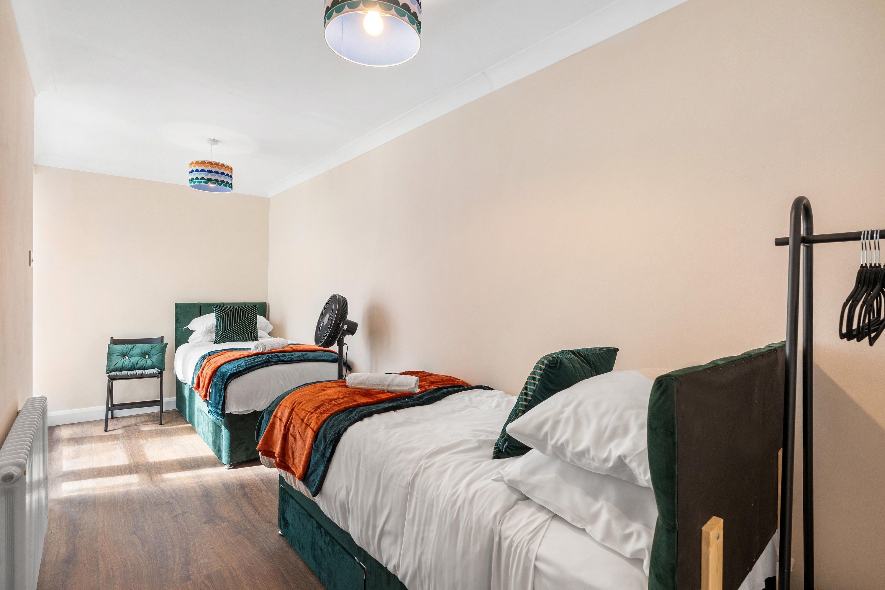A hotel room with two beds, a chair, a standing fan, and a coat rack.